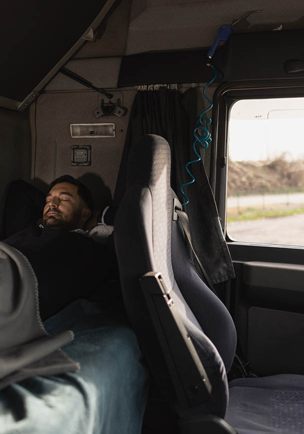 A person is resting with eyes closed on a bed inside a vehicle cabin next to a driver’s seat and a window showing an outdoor scene. Description generated by AI