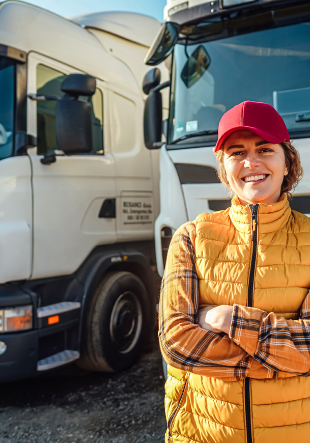 A person wearing a red cap and a yellow quilted vest is standing with arms crossed and smiling in front of two large white trucks. Description generated by AI