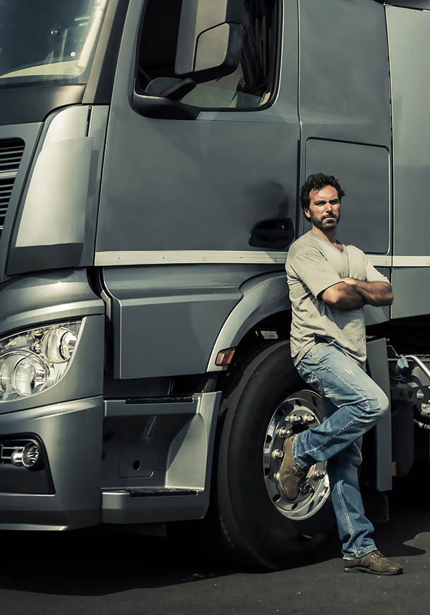 A person with curly hair wearing a light-colored shirt and jeans is standing with arms crossed and leaning against the front wheel of a large gray truck. Description generated by AI
