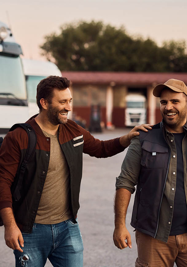 Two adults wearing casual outdoor clothing are smiling and interacting warmly near parked trucks in an outdoor setting. Description generated by AI