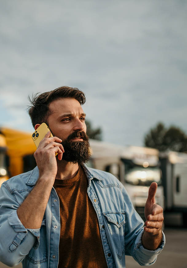 A person with a beard wearing a denim jacket and brown shirt is talking on a yellow phone outdoors with trucks in the background. Description generated by AI