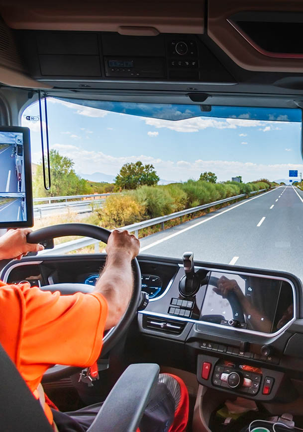 A person wearing an orange shirt is driving a large vehicle on a clear highway with green bushes and trees along the roadside under a blue sky with scattered clouds. Description generated by AI