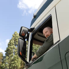 A person with short hair and a beard is leaning out of the window of a large green and white vehicle, smiling, with trees and a blue sky in the background. Description generated by AI