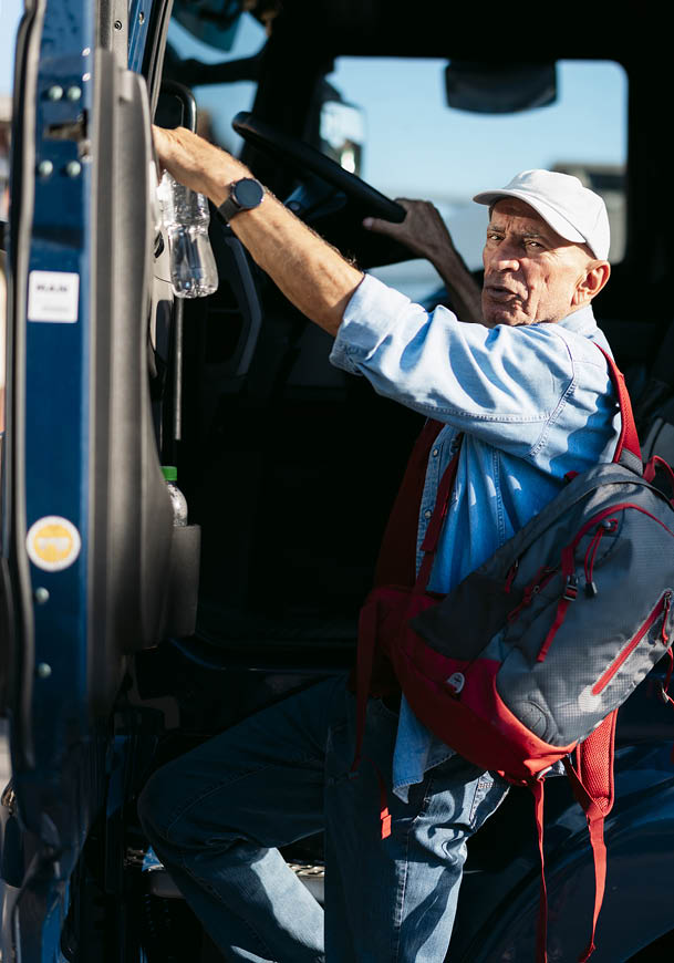 A senior wearing a light blue shirt, white cap, and backpack is climbing into a vehicle while holding a water bottle. Description generated by AI