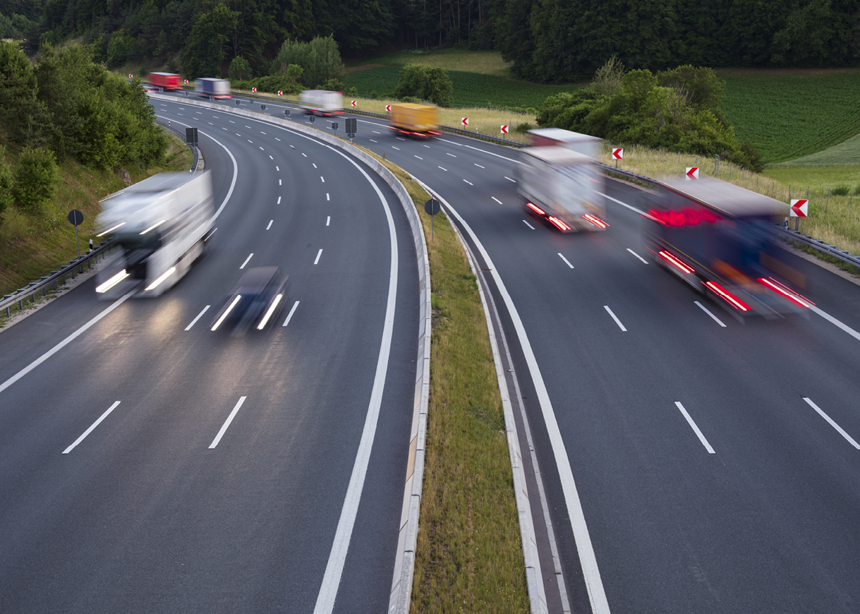 Multiple trucks are moving at high speed on a divided highway surrounded by greenery and trees. Description generated by AI