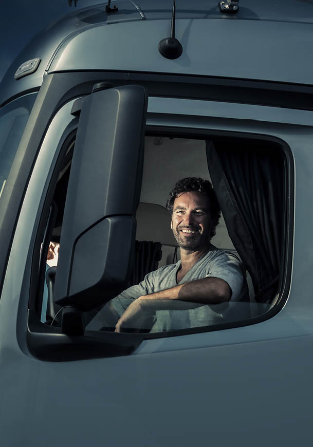 A person with curly hair and a beard is smiling while sitting inside the cab of a large vehicle, possibly a truck, with their arm resting on the open window. Description generated by AI