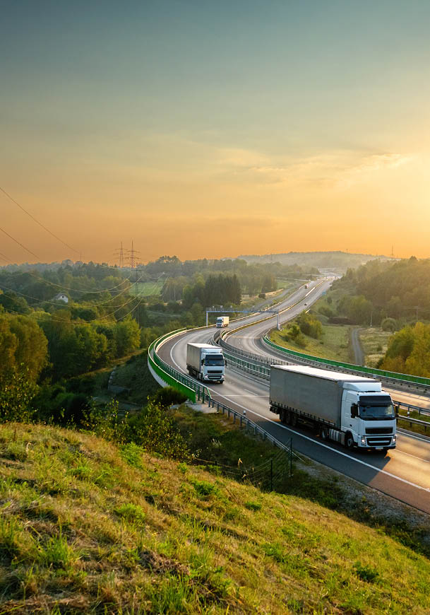 A winding highway stretches through a green, hilly landscape at sunset with several trucks driving along the road. Description generated by AI