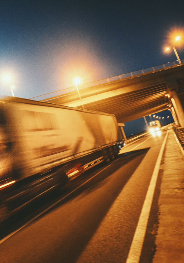 A large truck is driving quickly on a highway at night under an overpass with streetlights illuminating the scene. Description generated by AI