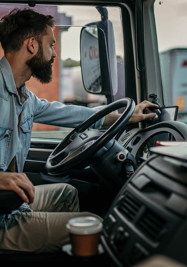 A person wearing a denim jacket and light-colored pants is sitting in the driver's seat of a car with a coffee cup placed near the dashboard. Description generated by AI