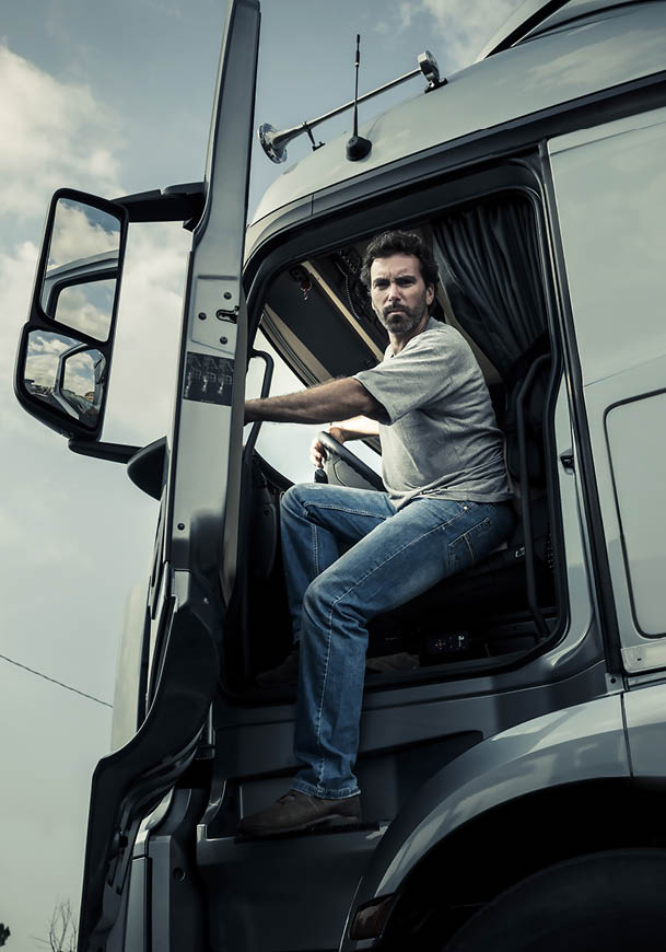 A person wearing a gray shirt and blue jeans is sitting in the driver's seat of a large truck with the door open, looking forward. Description generated by AI
