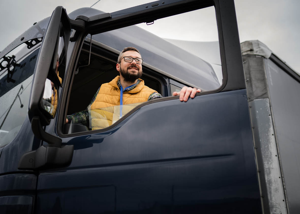 A person wearing glasses and a yellow vest is sitting in the driver's seat of a large blue truck with the door open, looking outside. Description generated by AI