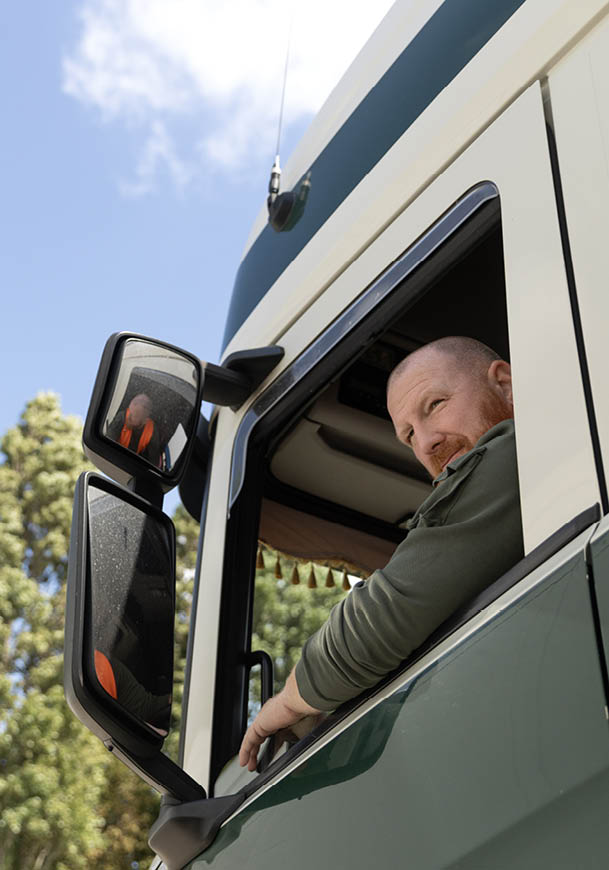 A person wearing a green long-sleeve shirt is leaning out of the window of a large green and white truck, looking outside. Description generated by AI